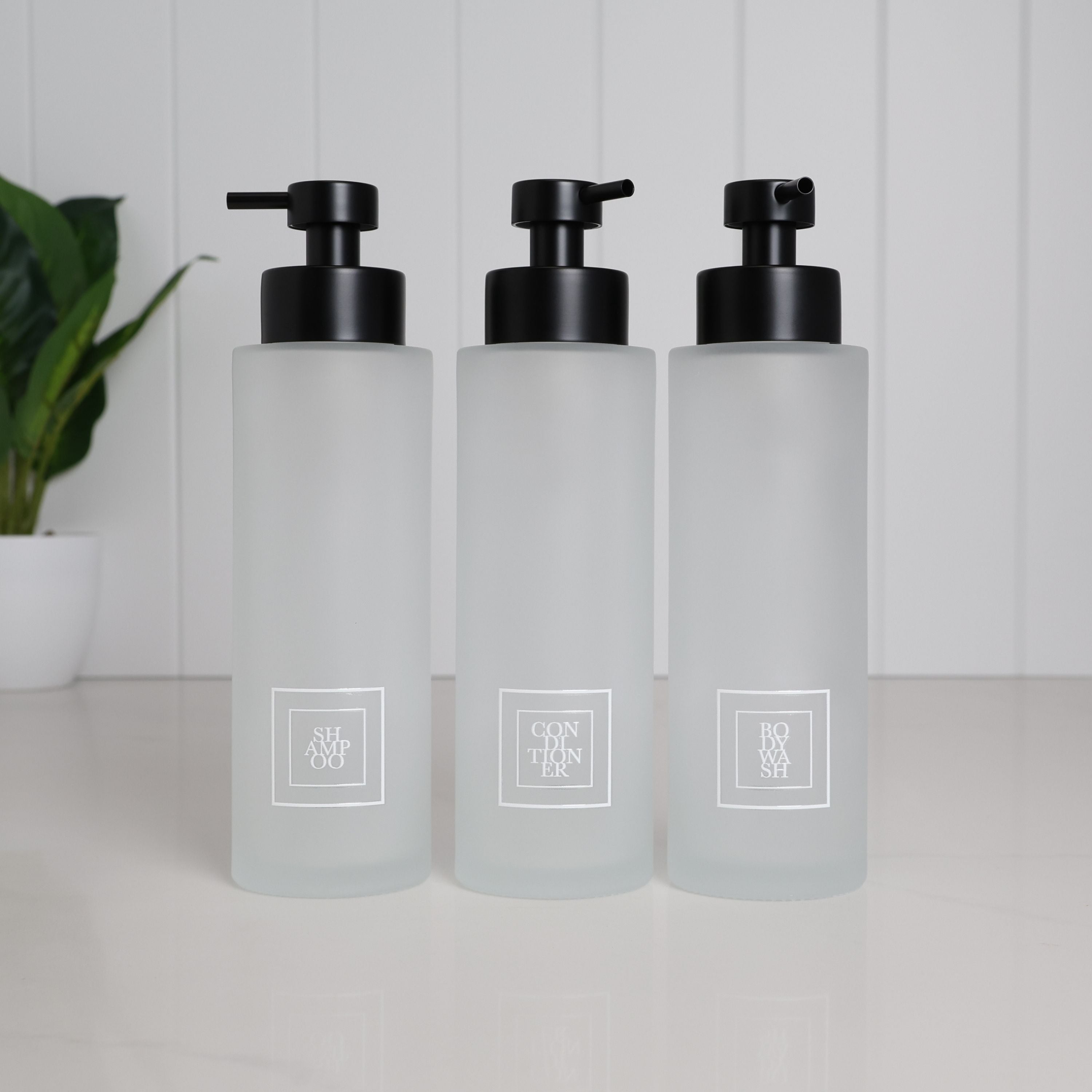 Set of three 500ml frosted glass soap dispensers with white shampoo, conditioner and body wash labels and satin black pumps, designed as refillable bathroom bottles for a modern shower or countertop.