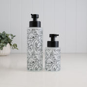 Two frosted glass pump bottles with black caps, decorated with black line art illustrations, standing on a white surface against a white paneled background.