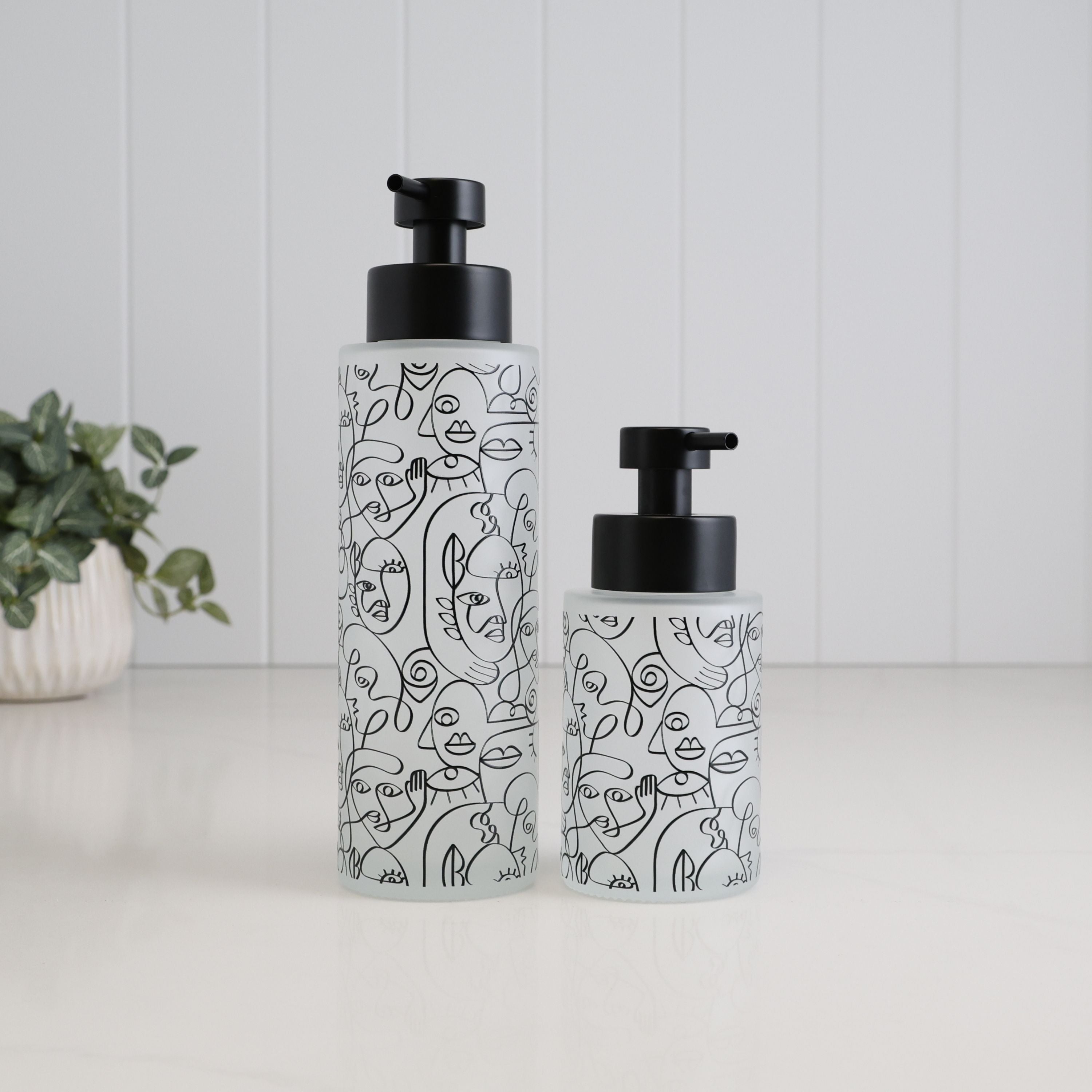 Two frosted glass pump bottles with black caps, decorated with black line art illustrations, standing on a white surface against a white paneled background.