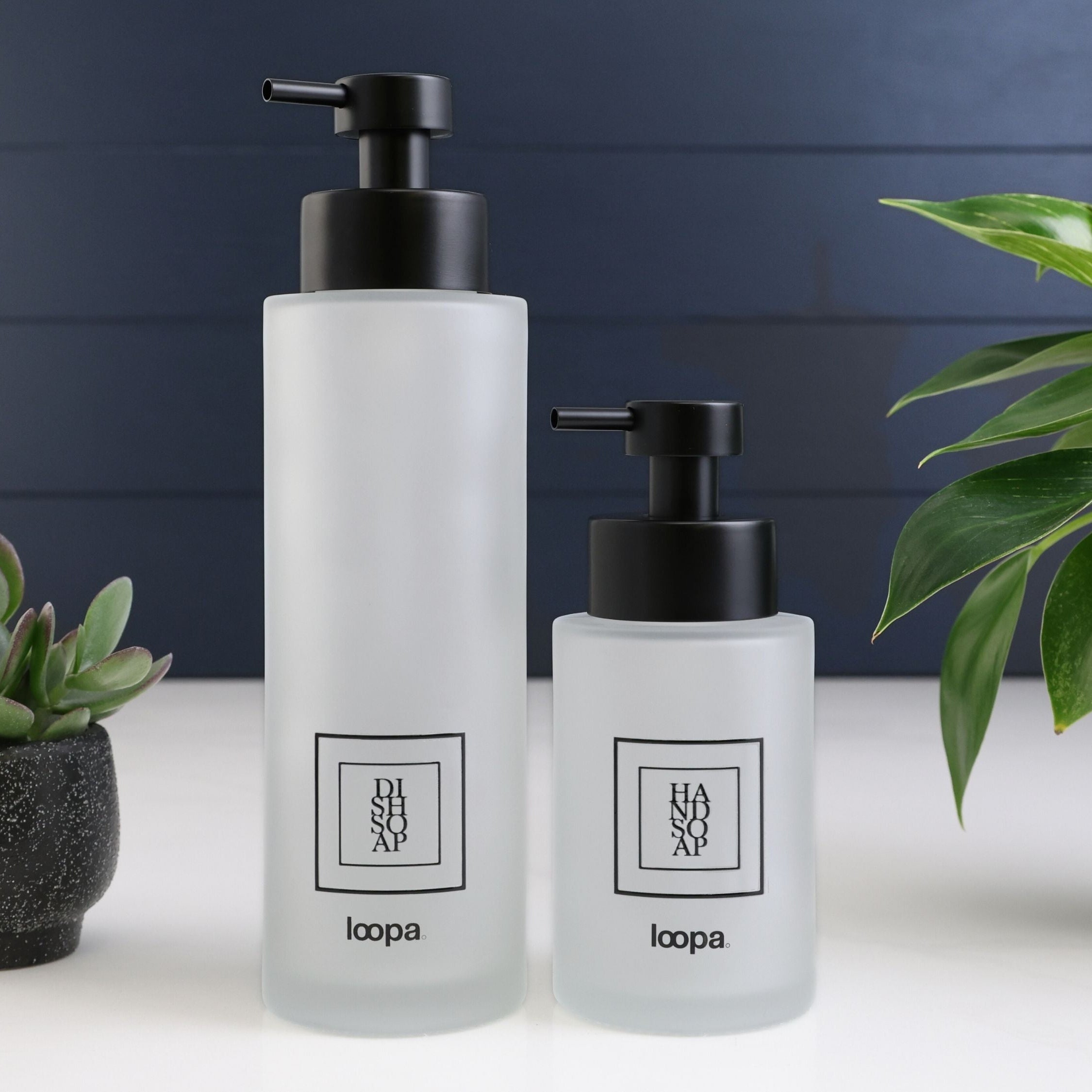 Square frosted glass bottle dispenser with a black label, presented as a refillable option for modern kitchen or bathroom use