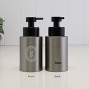 Two brushed stainless steel pump bottles shown side by side on a white surface against a white paneled wall. The left bottle shows the front with a black pump and a circular silver logo reading HADSOAP, and the right bottle shows the back with a black pump and the word loopa.
