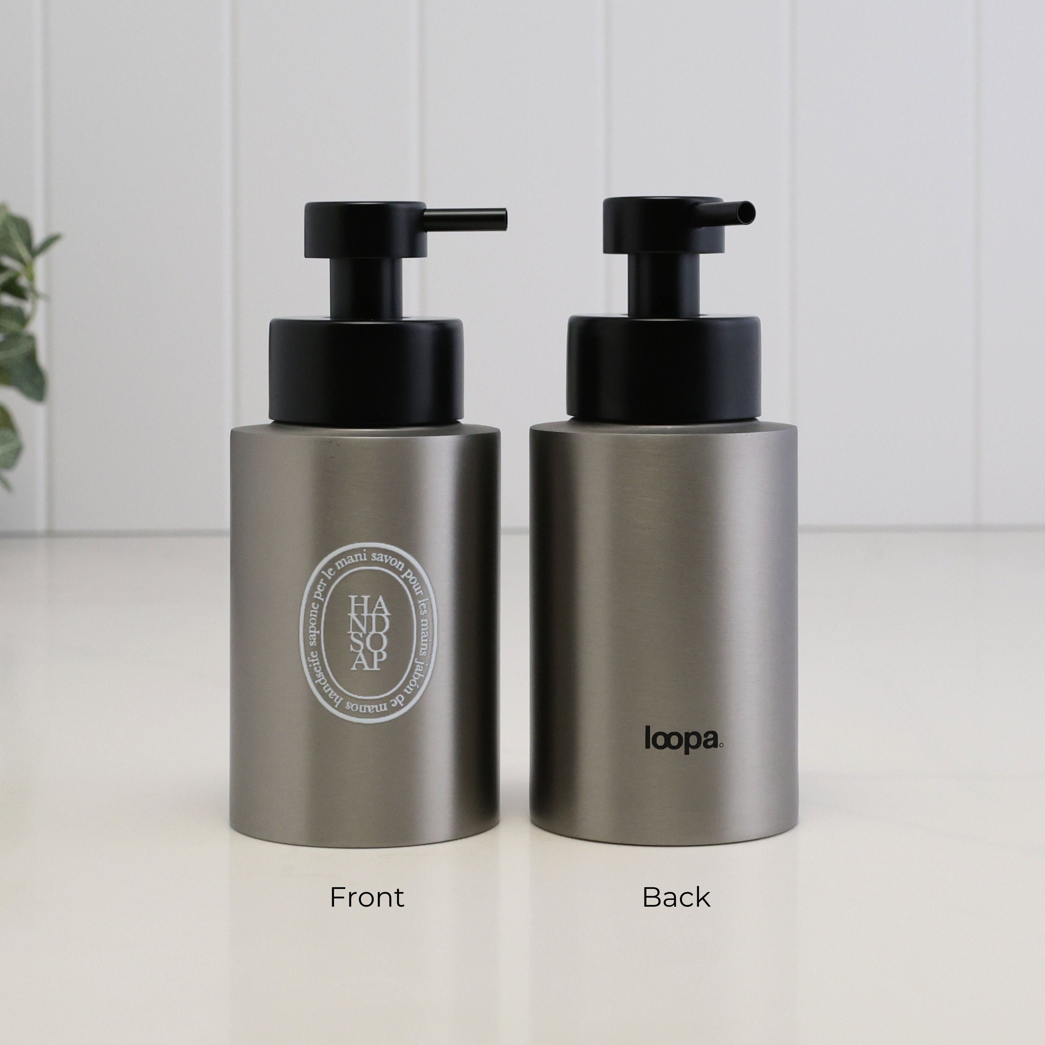 Two brushed stainless steel pump bottles shown side by side on a white surface against a white paneled wall. The left bottle shows the front with a black pump and a circular silver logo reading HADSOAP, and the right bottle shows the back with a black pump and the word loopa.