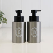 Two metallic silver Diptyque inspired Hand Soap bottles with black pumps, standing side by side on a white surface against a white paneled background.