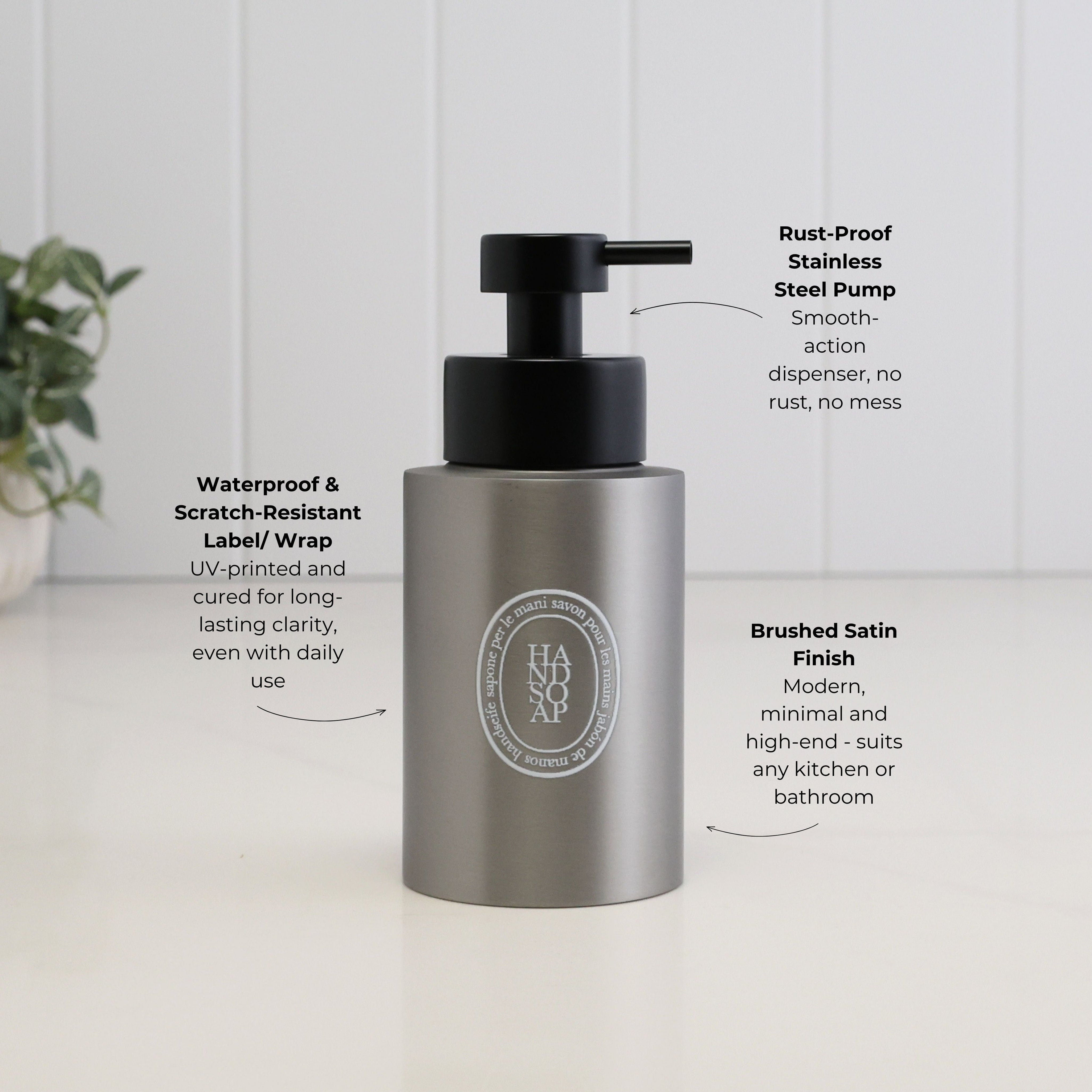 A stainless steel pump bottle with a black dispenser, shown on a white surface with white paneled walls in the background. Text callouts describe features such as rust-proof stainless steel pump, waterproof and scratch-resistant label/wrap, and brushed satin finish.