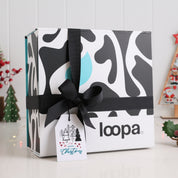 Loopa Christmas gift box with black ribbon and festive tag, displayed on white surface with holiday decorations in background.
