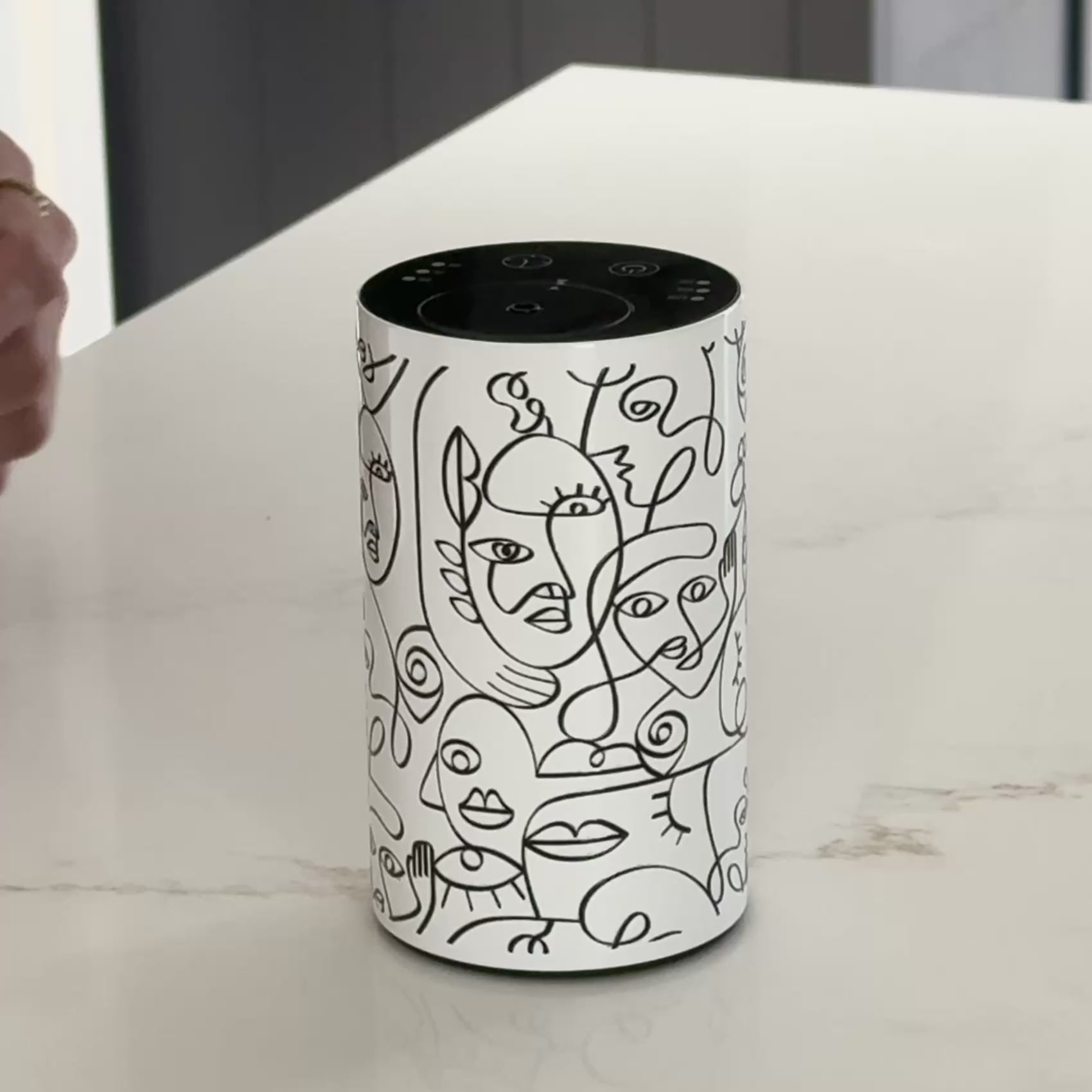 Video showcasing the Loopa white waterless essential oil diffuser with black abstract face illustrations and a minimalist cylindrical metal body. The close-up highlights the decorative line art design and top control panel for adjusting fragrance intensity and timer settings. This cordless, rechargeable aromatherapy diffuser uses pure essential oils without water or heat, offering portable home fragrance for bedrooms, bathrooms and living spaces.