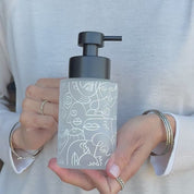 Frosted glass soap dispenser with white abstract line art design and satin black pump, shown held in hand to demonstrate the size and decorative finish of the refillable bathroom bottle.