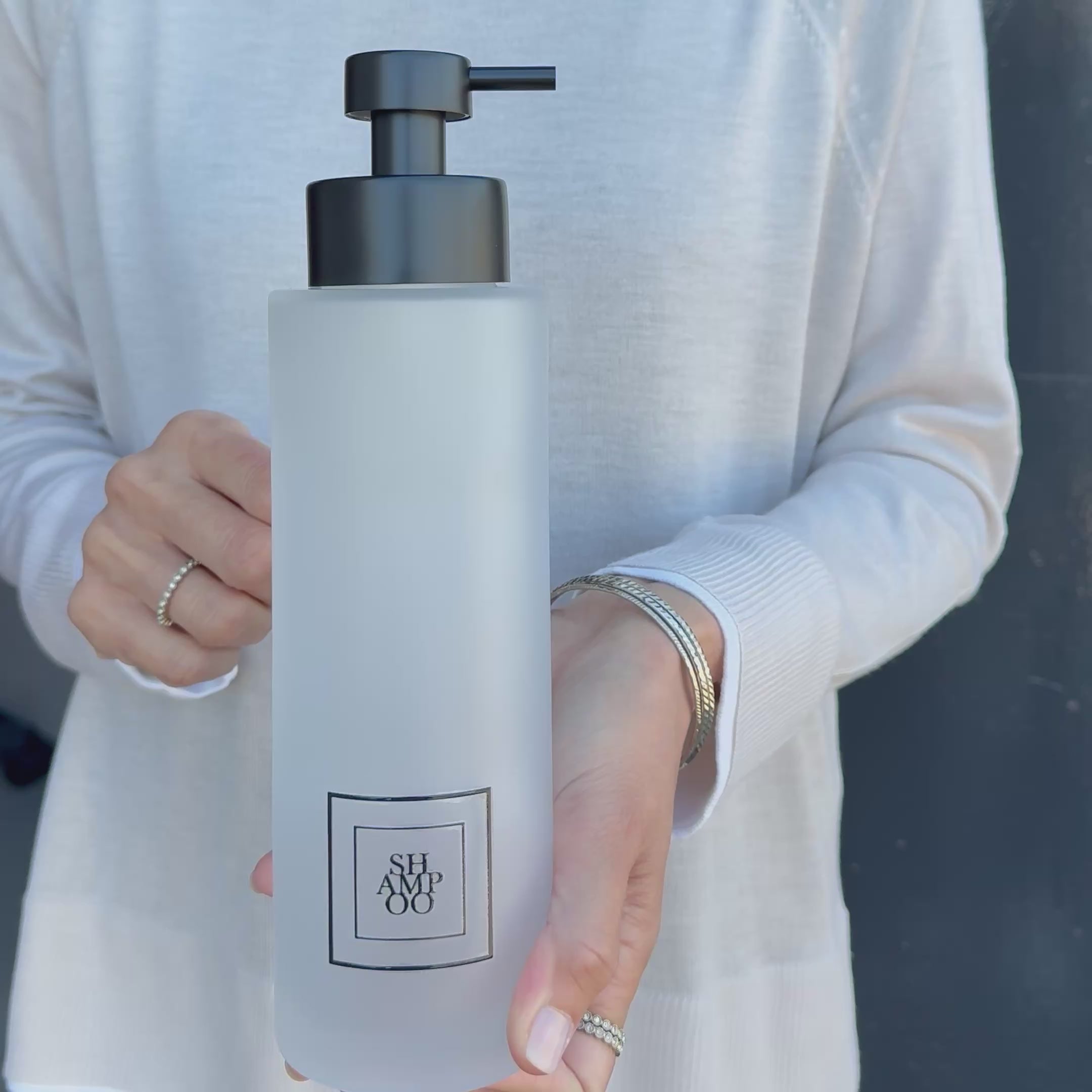 500ml frosted glass shampoo dispenser with black square label and satin black stainless steel pump, shown held in hand to demonstrate the tall refillable bottle size and premium bathroom design.