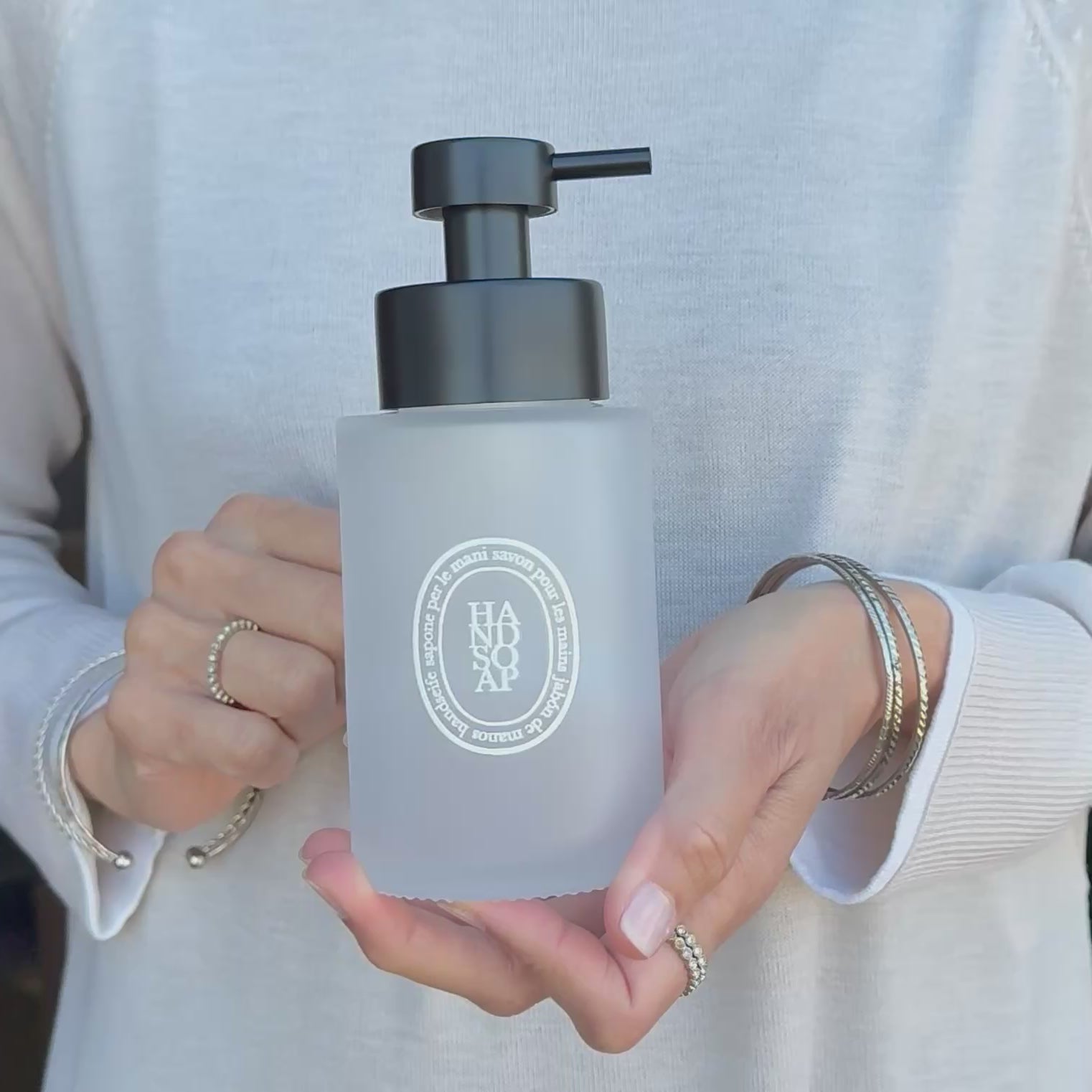 Video showing a 250ml frosted glass hand soap dispenser with white oval label and satin black stainless steel pump, held in hand to demonstrate its compact size and frosted finish.