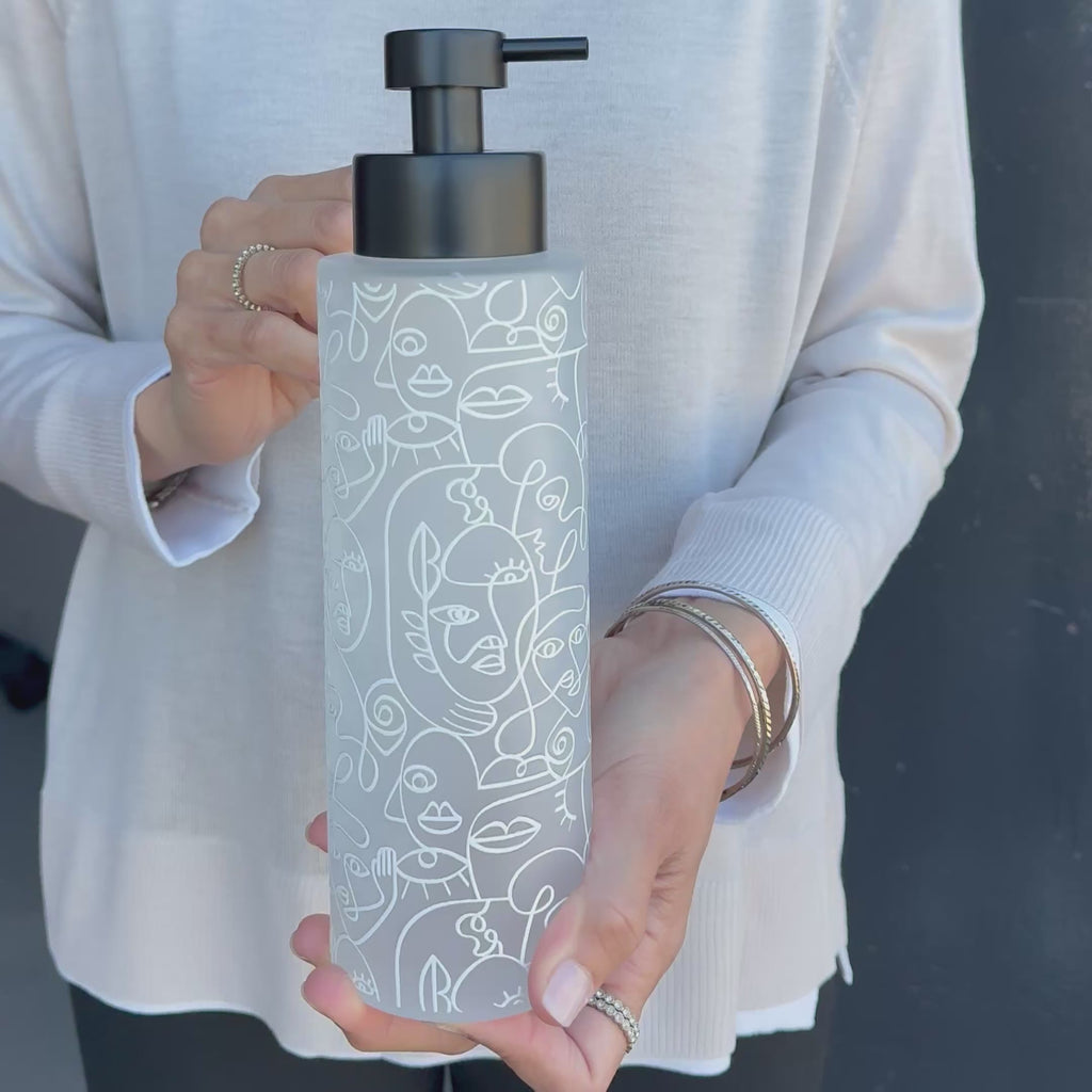 Frosted glass 500ml soap dispenser with white abstract face line art and satin black pump, shown held in hand to demonstrate the tall bottle size, decorative wrap and modern refillable design.