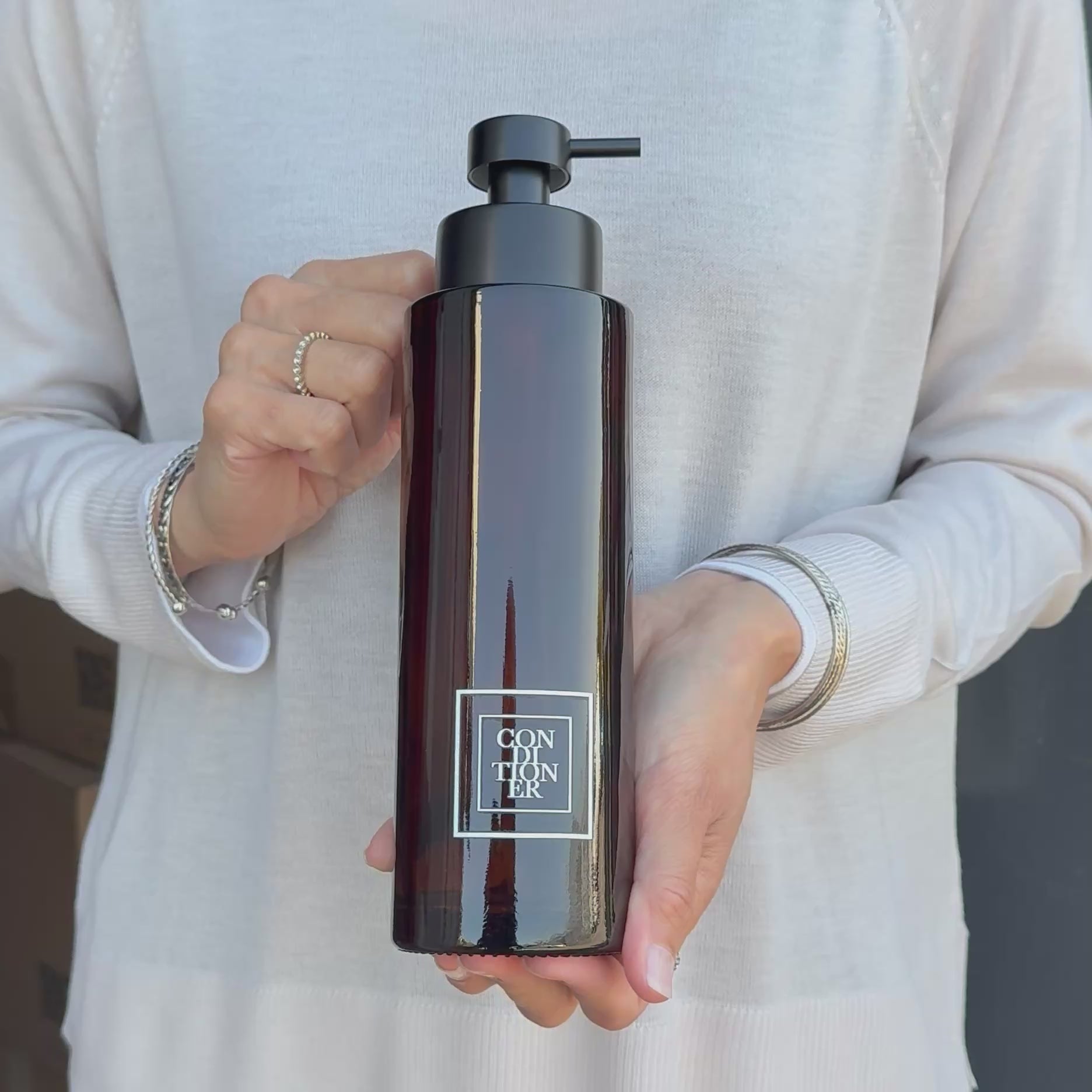 Video showing a 500ml amber glass conditioner dispenser with a white square label and satin black pump, held to show the bottle’s size and design.