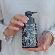 Video showing an amber glass soap dispenser with white abstract face line art wrap and satin black pump, held in hand to show the design and size.