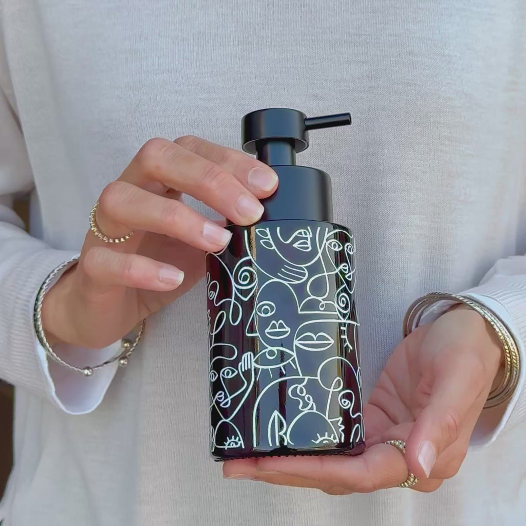 Video showing an amber glass soap dispenser with white abstract face line art wrap and satin black pump, held in hand to show the design and size.