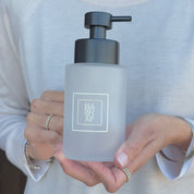 250ml frosted glass hand soap dispenser with white square Hand Soap label and satin black pump, shown held in hand to demonstrate the compact size of the refillable bathroom sink bottle.
