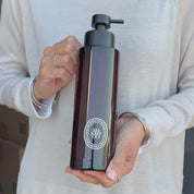 Video showing a 500ml amber glass shampoo dispenser with a white round label and satin black stainless steel pump, held to demonstrate its size, finish and design.