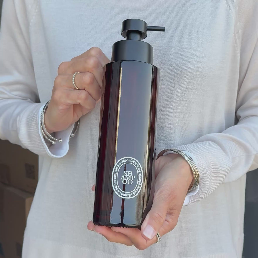 Video showing a 500ml amber glass shampoo dispenser with a white round label and satin black stainless steel pump, held to demonstrate its size, finish and design.