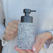 250ml frosted glass soap dispenser with black abstract face line art and satin black pump, shown held in hand to demonstrate the compact size and decorative refillable design for hand soap or lotion.