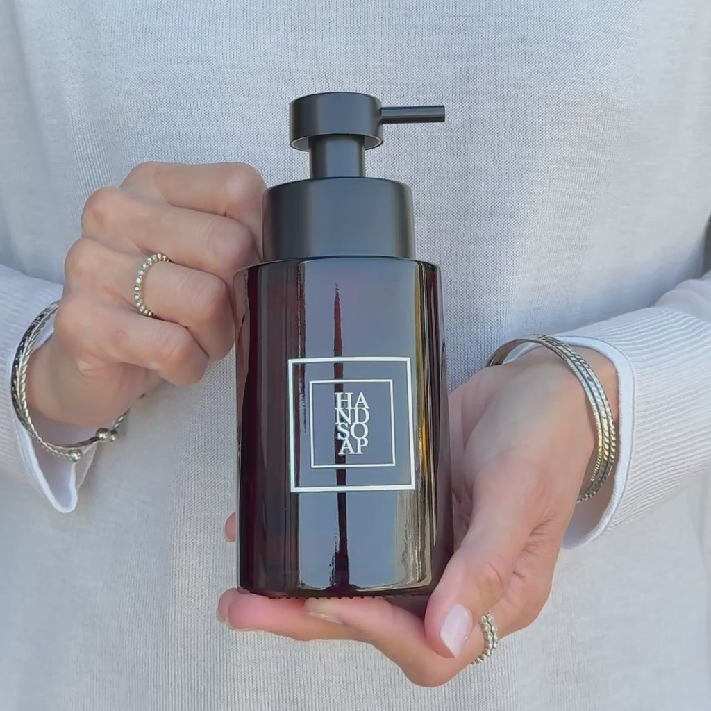 Video showing a 250ml amber glass hand soap dispenser with a white square label and satin black pump, held in the hands to demonstrate its compact size and design.