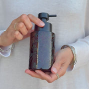 Video showing an amber glass soap dispenser with subtle black abstract face line art wrap and satin black pump, held in hand to show the decorative design and size.