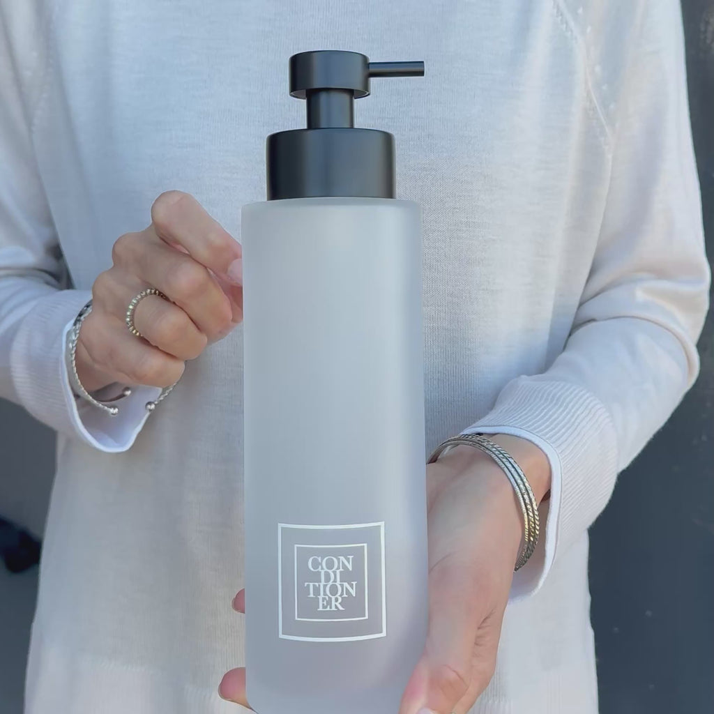 500ml frosted glass conditioner dispenser with white square Conditioner label and satin black pump, shown held in hand to demonstrate the size and premium refillable bathroom bottle design.