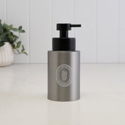 A matte gray cylindrical pump bottle with a black dispenser, featuring a circular silver Diptyque logo on the front, set on a white surface with a white paneled background and a blurred green plant in the corner.