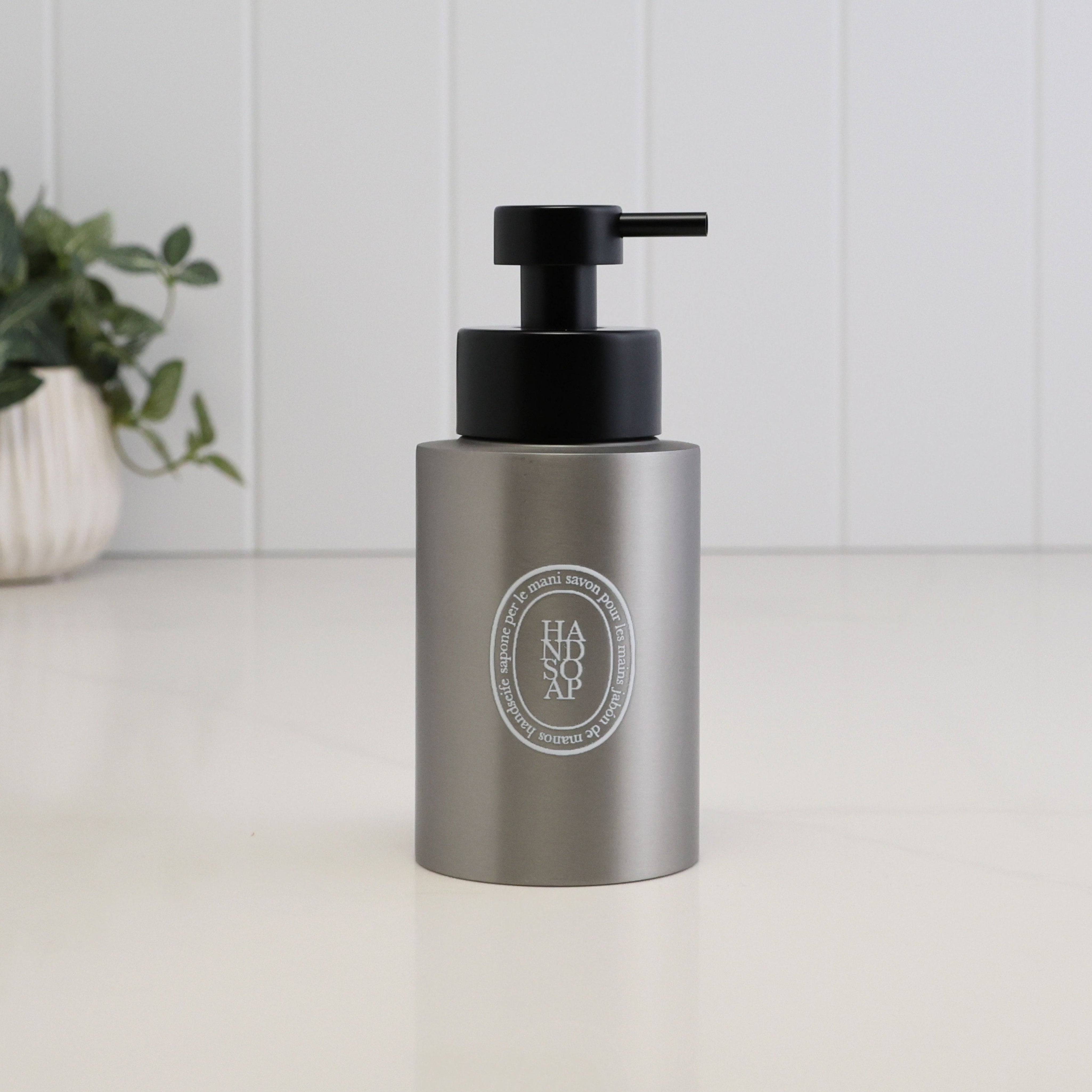 A matte gray cylindrical pump bottle with a black dispenser, featuring a circular silver Diptyque logo on the front, set on a white surface with a white paneled background and a blurred green plant in the corner.