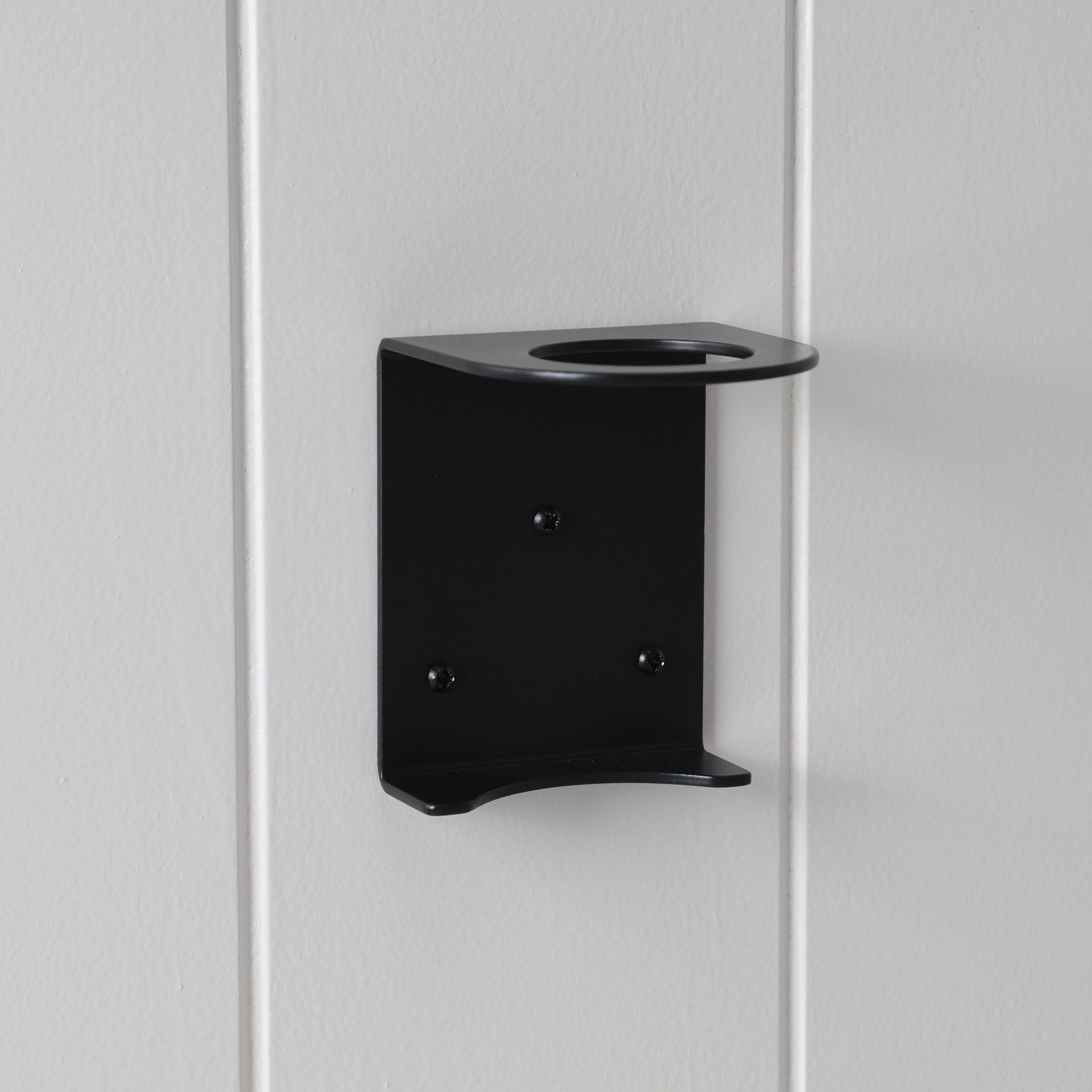 Loopa single wall-mounted soap dispenser bracket in black powder-coated aluminium, securely fixed to a white panelled wall. Featuring a circular bottle opening and supporting base, this minimalist holder is designed for Loopa 250ml and 500ml refillable soap dispenser bottles. Ideal for modern bathrooms, showers and kitchens.