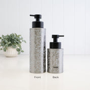 Two metallic silver pump bottles with black dispensers, shown from front and back on a white surface against a white paneled wall. The bottles have a white line-art pattern and the brand name 'loopa' is visible on the back bottle.