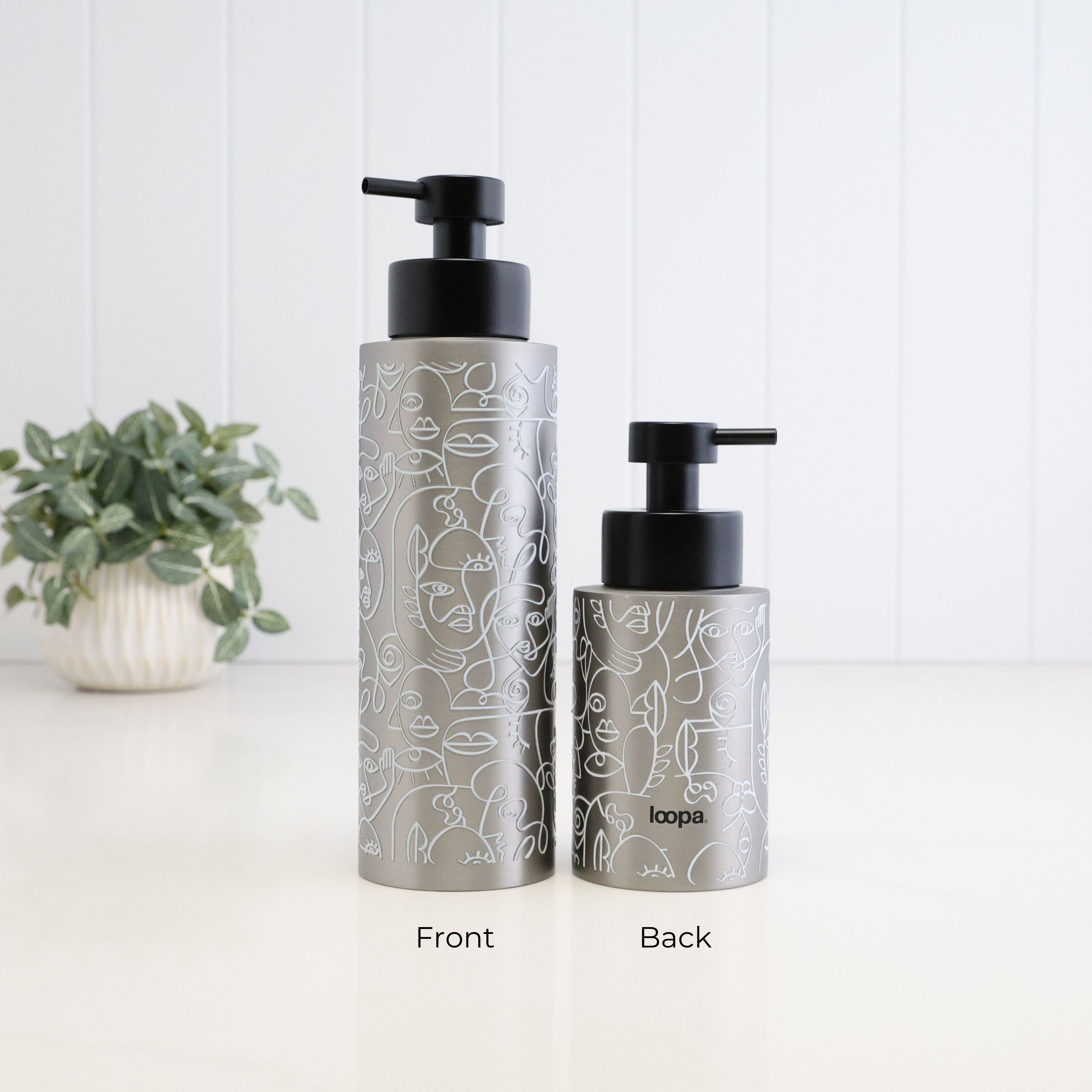 Two metallic silver pump bottles with black dispensers, shown from front and back on a white surface against a white paneled wall. The bottles have a white line-art pattern and the brand name 'loopa' is visible on the back bottle.