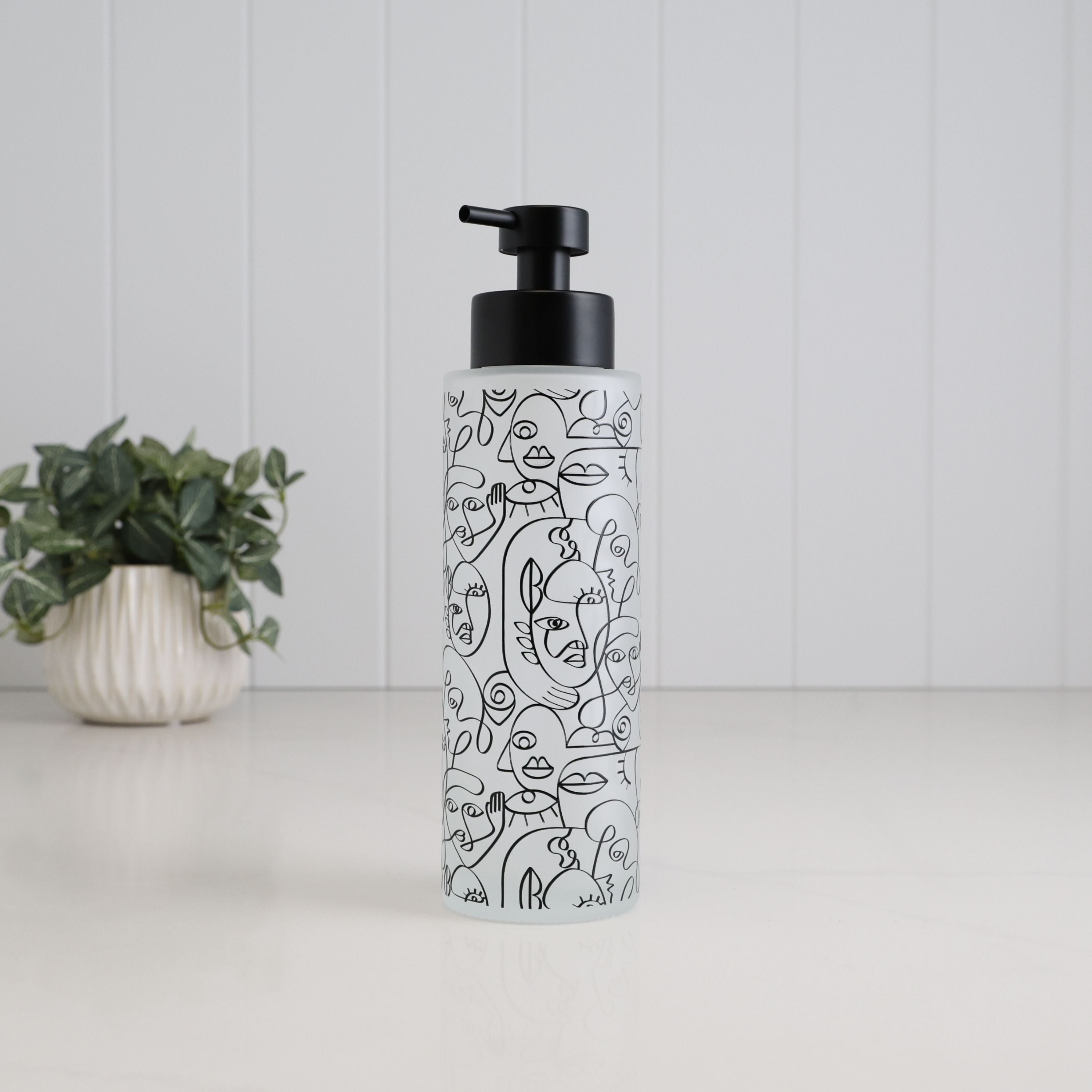 Tall frosted glass soap dispenser with black abstract face line art and satin black pump, a refillable 500ml bottle designed for hand soap, shampoo, conditioner or body wash in modern bathrooms.