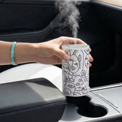 Loopa white waterless essential oil diffuser with black abstract face line art, held above a car cup holder while releasing a fine fragrance mist. This cordless, rechargeable car aroma diffuser features a cylindrical metal body and uses essential oils directly without water or heat. A portable aromatherapy diffuser designed for convenient fragrance on car journeys.