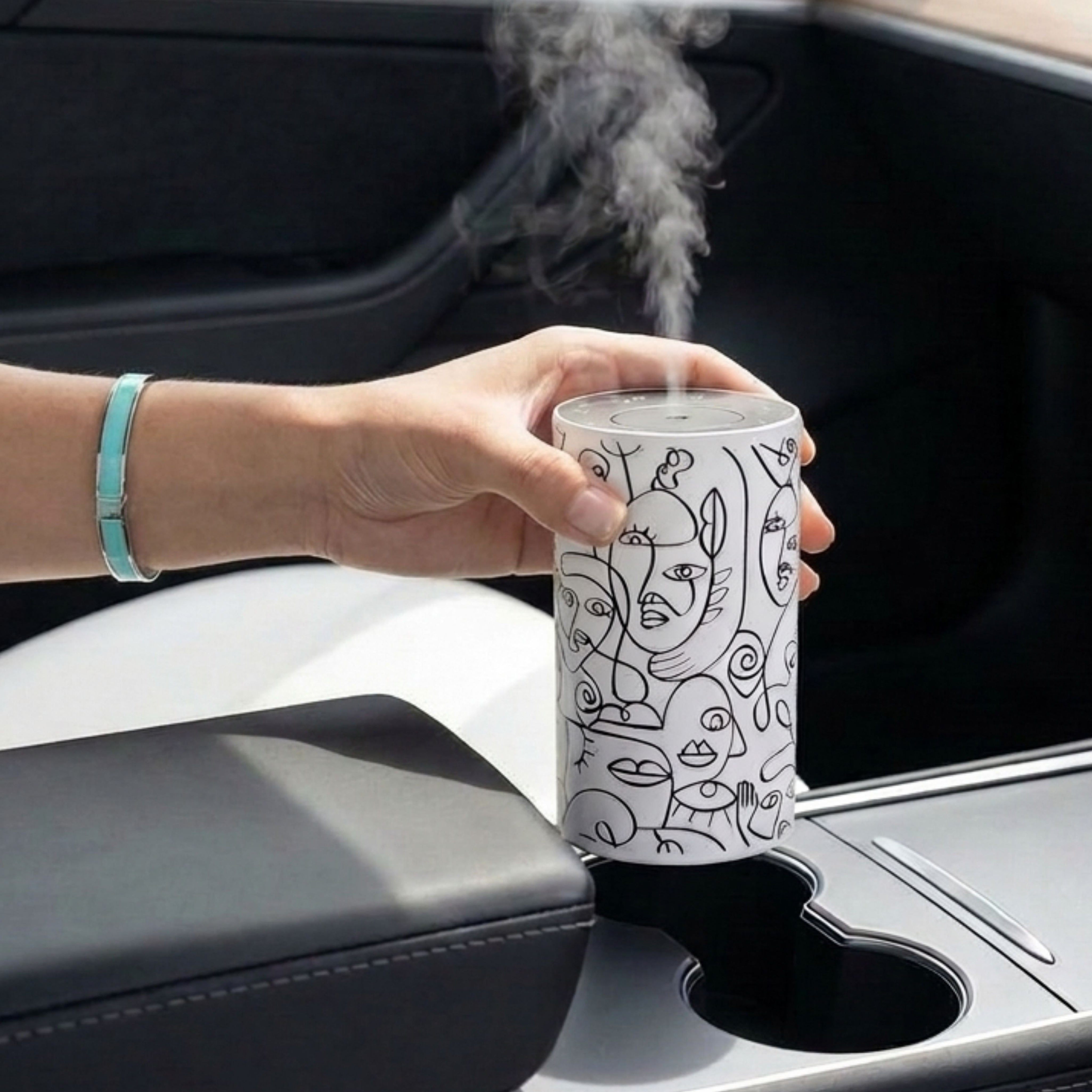 Loopa white waterless essential oil diffuser with black abstract face line art, held above a car cup holder while releasing a fine fragrance mist. This cordless, rechargeable car aroma diffuser features a cylindrical metal body and uses essential oils directly without water or heat. A portable aromatherapy diffuser designed for convenient fragrance on car journeys.