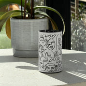Loopa white waterless essential oil diffuser with black abstract face line art, displayed on a sunlit windowsill beside a large potted plant. The minimalist cylindrical metal diffuser features a black control panel and cordless rechargeable design, blending into contemporary home interiors. Ideal for adding essential oil fragrance to living rooms, bedrooms and home offices without water or heat.