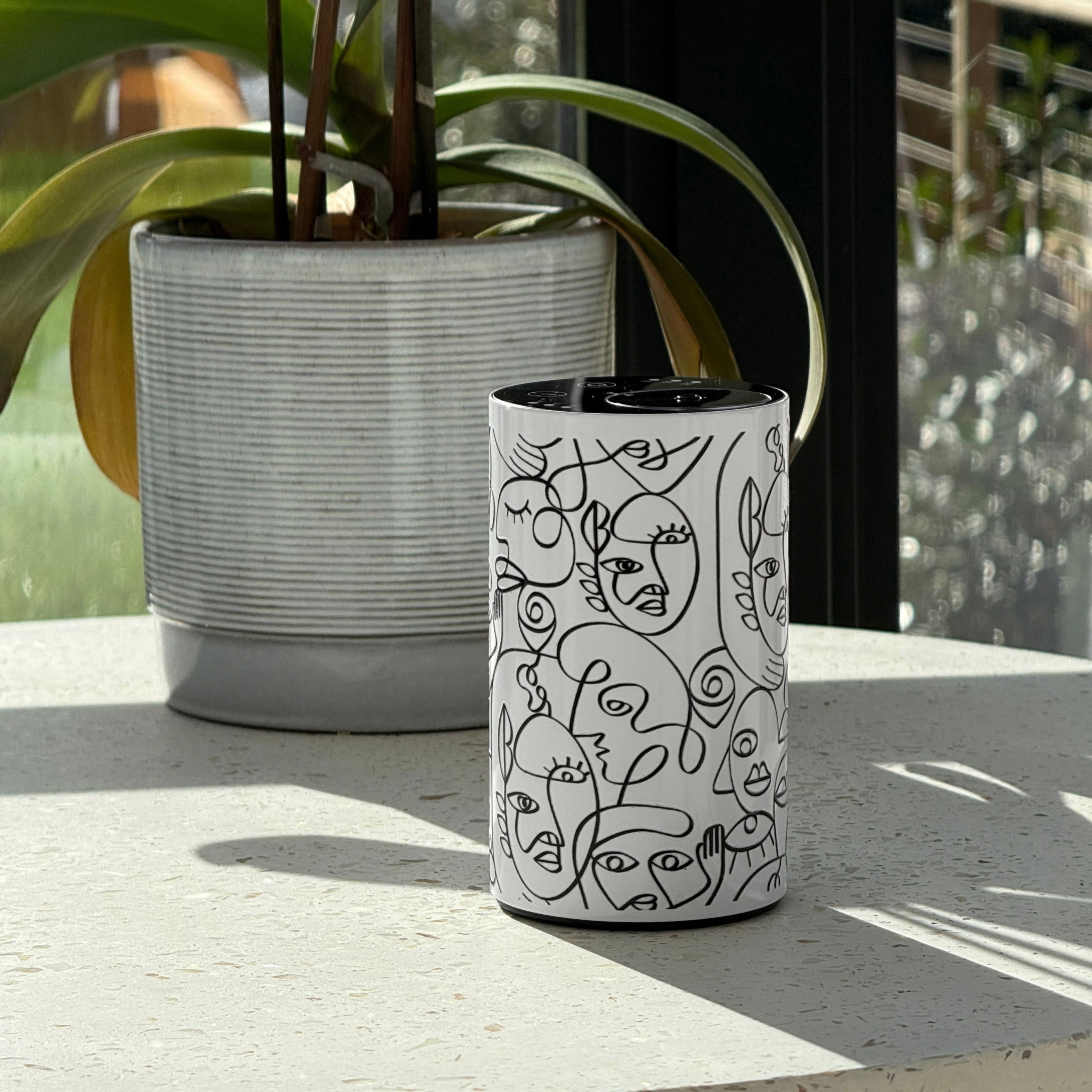 Loopa white waterless essential oil diffuser with black abstract face line art, displayed on a sunlit windowsill beside a large potted plant. The minimalist cylindrical metal diffuser features a black control panel and cordless rechargeable design, blending into contemporary home interiors. Ideal for adding essential oil fragrance to living rooms, bedrooms and home offices without water or heat.