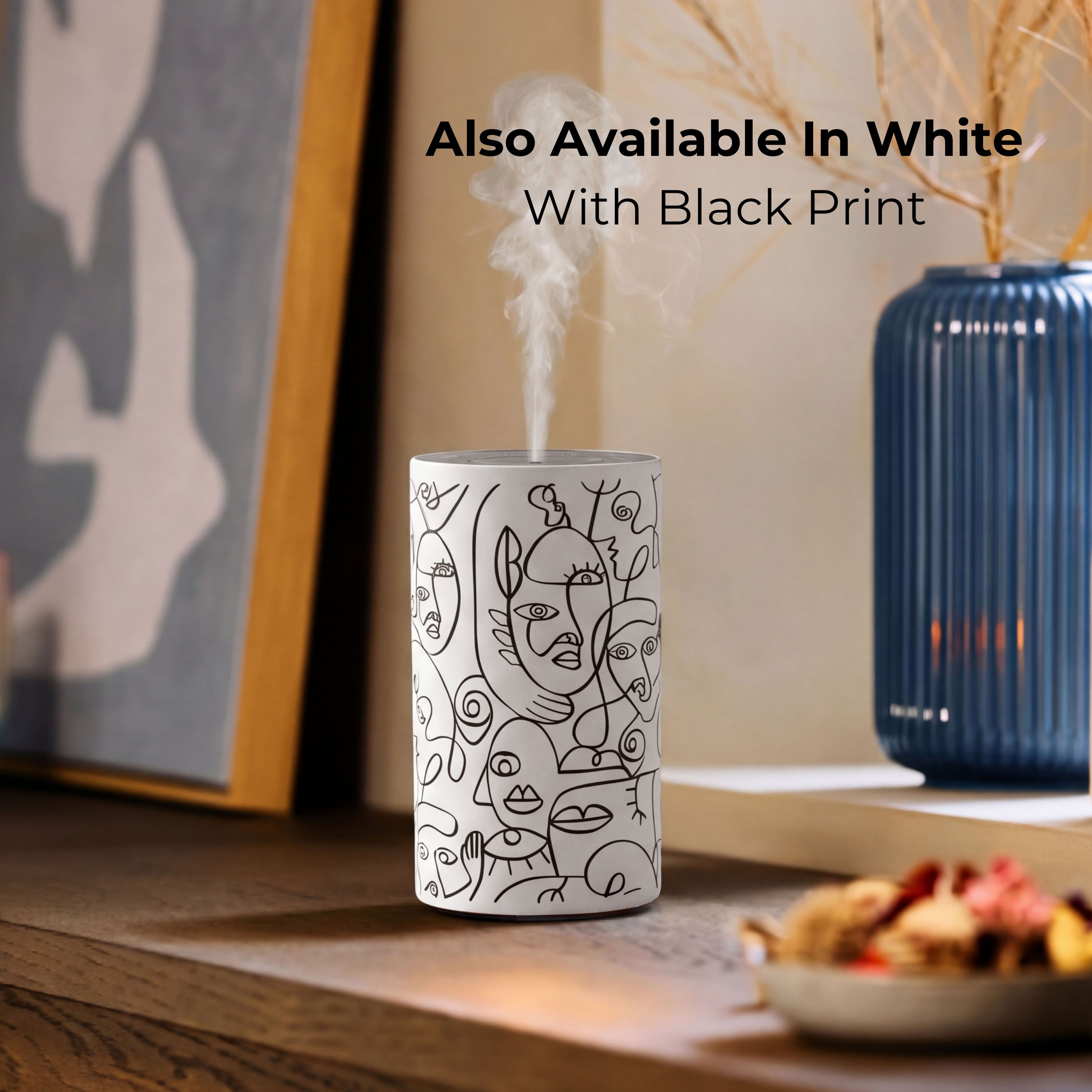Loopa white cordless waterless essential oil diffuser with black abstract line art face design, releasing a fine fragrance mist on a wooden table. Styled beside framed artwork and a blue ceramic vase, this rechargeable portable aromatherapy diffuser uses essential oils directly without water. Alternative white colour option for modern bedrooms, living rooms and home interiors.