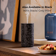 Black Loopa waterless essential oil diffuser with grey abstract face line art, shown releasing fragrance mist on a wooden sideboard. This alternative colour option features a cylindrical metal body with a rechargeable cordless design. Styled alongside framed artwork and a blue decorative vase, it offers a contemporary home fragrance solution for bedrooms, living rooms and offices.