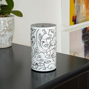Loopa white waterless essential oil diffuser featuring black abstract face illustrations on a cylindrical metal body. Displayed on a black cabinet alongside a decorative plant and colourful artwork, this cordless rechargeable aroma diffuser complements modern home interiors. Designed for bedrooms, living rooms and offices, it disperses pure essential oils without water or heat.