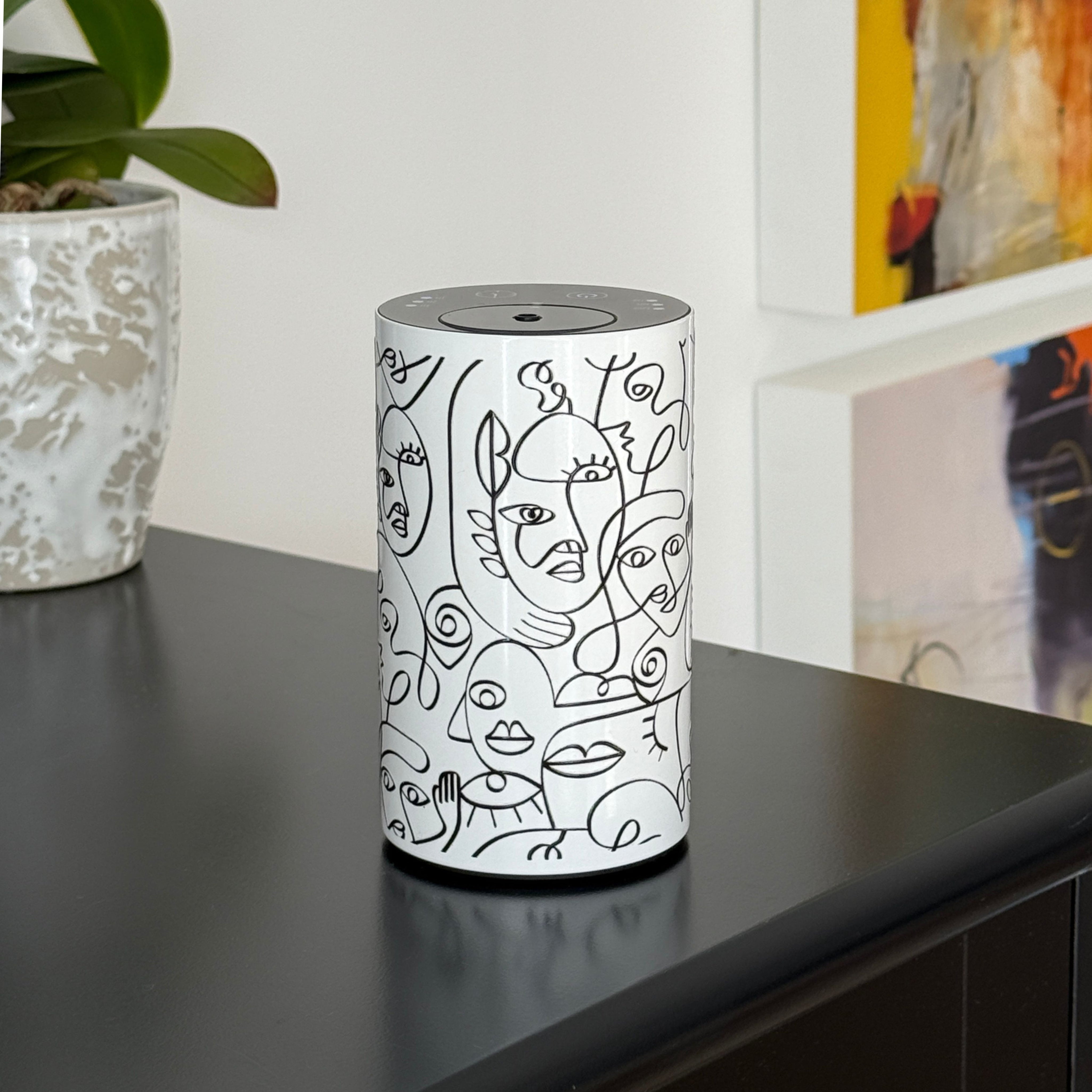 Loopa white waterless essential oil diffuser featuring black abstract face illustrations on a cylindrical metal body. Displayed on a black cabinet alongside a decorative plant and colourful artwork, this cordless rechargeable aroma diffuser complements modern home interiors. Designed for bedrooms, living rooms and offices, it disperses pure essential oils without water or heat.