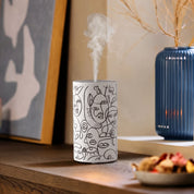 Loopa white rechargeable waterless essential oil diffuser with black abstract face line art design, releasing a fine fragrance mist on a wooden table. Styled alongside framed artwork and a blue ceramic vase, this cordless metal aromatherapy diffuser uses pure essential oils without water or heat. A contemporary home fragrance accessory for bedrooms, living rooms and offices.