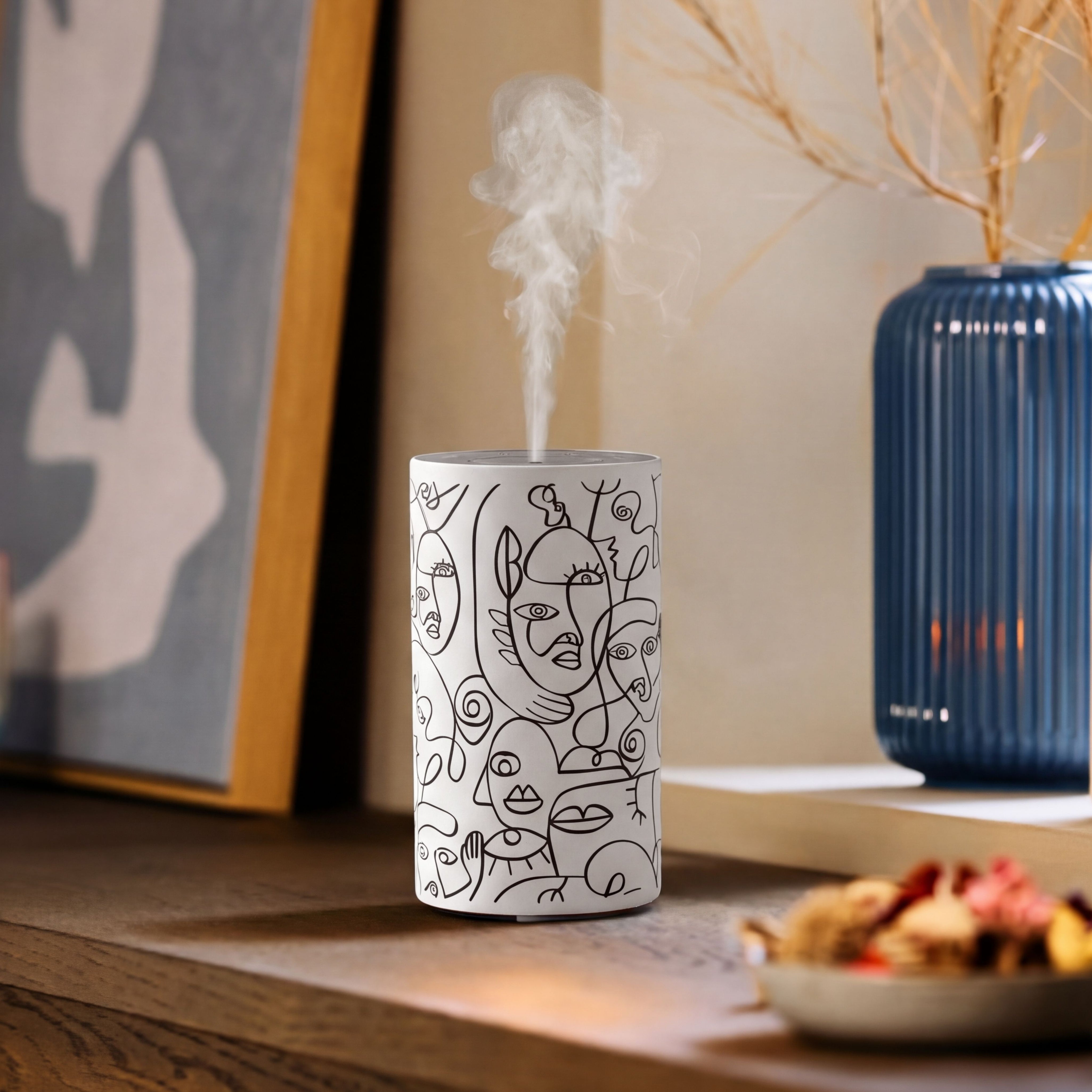 Loopa white rechargeable waterless essential oil diffuser with black abstract face line art design, releasing a fine fragrance mist on a wooden table. Styled alongside framed artwork and a blue ceramic vase, this cordless metal aromatherapy diffuser uses pure essential oils without water or heat. A contemporary home fragrance accessory for bedrooms, living rooms and offices.